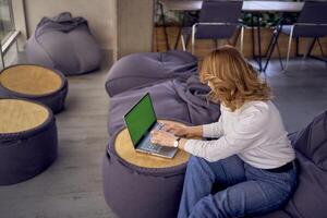 een vrouw in gewoontjes kleren is zittend Bij een laptop Aan Boon zak in voorkant van een panoramisch venster met een visie van de stad, groen scherm, chroma sleutel foto