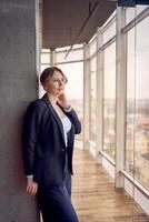 aantrekkelijk midden- leeftijd vrouw in jasje en wit t-shirt poseren in modern kantoor met panoramisch ramen foto