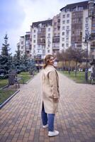 jong volwassen vrouw in jeans en zand jas wandelingen door de stad Aan een voorjaar dag, modern architectuur achter foto