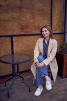 jong volwassen vrouw in jeans en zand jas met grunge achtergrond, roestig muur met koffie belettering foto