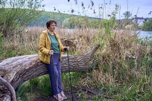 ouderen vrouw rust Aan de rivier- bank na nordic wandelen oefening foto