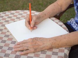 detailopname van senior vrouw gebruik makend van potlood schrijven een boek Bij de tafel terwijl zittend Aan een stoel in een tuin. ruimte voor tekst. concept van oud mensen en ontspanning foto