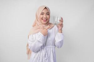 gelukkig jong Aziatisch moslim vrouw in een sluier hijab richten naar een glas van Doorzichtig water ze houdt. Ramadan advertentie concept foto