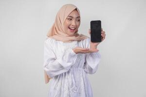 portret van jong Aziatisch moslim vrouw vervelend wit jurk en hijab glimlachen gelukkig, richten naar de blanco scherm kopiëren ruimte Aan haar telefoon en tonen scherm naar de camera foto