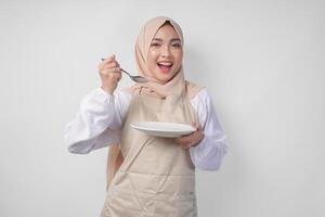 hongerig jong Aziatisch moslim vrouw in hijab en room schort Holding lepel en aan het eten van een leeg bord met kopiëren ruimte over- het. Ramadan concept foto