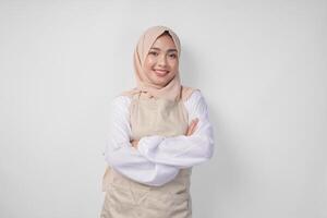 jong Aziatisch moslim vrouw vervelend hijab en room schort glimlachen naar de camera met gekruiste armen houding vol van vertrouwen foto