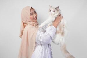 portret van een mooi jong moslim vrouw vervelend wit jurk en hijaab, glimlachen en Holding een wit lappenpop kat over- geïsoleerd wit achtergrond foto
