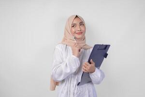 attent jong Aziatisch moslim vrouw vervelend wit jurk en hijab Holding een document, denken terwijl op zoek naar de kopiëren ruimte bovenstaande, geïsoleerd door wit achtergrond. Ramadan en eid mubarak concept foto