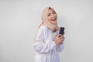 opgewonden Aziatisch moslim vrouw vervelend wit jurk en hijab Holding haar smartphone terwijl glimlachen vrolijk, geïsoleerd door wit achtergrond. Ramadhan en eid mubarak concept foto
