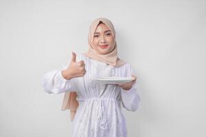 opgewonden jong Aziatisch moslim vrouw in hijab presenteren leeg wit bord kopiëren ruimte terwijl aan het doen duimen omhoog gebaar foto