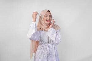 opgewonden jong Aziatisch moslim vrouw vervelend wit jurk en hijab testen of toepassen huid zorg serum Aan gezicht. gelaats en schoonheid concept foto