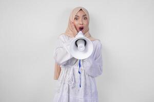 dolblij Aziatisch vrouw vervelend hijab en wit jurk geschreeuw naar de megafoon maken Aankondiging, staand over- geïsoleerd wit achtergrond foto