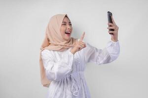 jong Aziatisch moslim vrouw met een gelukkig geslaagd uitdrukking vervelend wit jurk en hijab Holding smartphone, geïsoleerd door wit achtergrond. Ramadan en eid mubarak concept foto