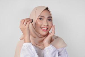 opgewonden jong Aziatisch moslim vrouw vervelend wit jurk en hijab testen of toepassen huid zorg serum Aan gezicht. gelaats en schoonheid concept foto