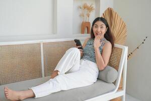 geschokt Aziatisch vrouw Holding en zien telefoon met mond breed Open terwijl houdende Aan een sofa in een villa voor vakantie foto