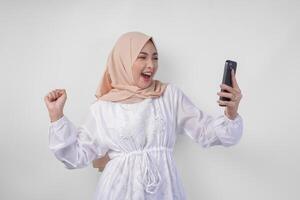 jong Aziatisch moslim vrouw met een gelukkig geslaagd uitdrukking vervelend wit jurk en hijab Holding smartphone, geïsoleerd door wit achtergrond. Ramadan en eid mubarak concept foto