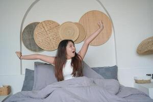 slaperig Aziatisch jong vrouw geeuwen en verhogen hand- ontwaken omhoog in vroeg ochtend- na diep slaap, vredig ochtend- vakantie concept foto
