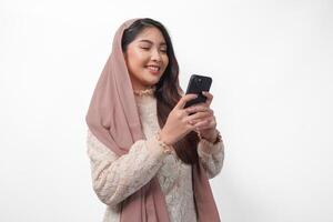 vrolijk jong Aziatisch moslim vrouw in sluier hijab glimlachen en gevoel gelukkig terwijl Holding een smartphone van kant visie. Ramadan en eid mubarak concept foto