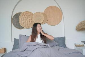 slaperig Aziatisch jong vrouw geeuwen en verhogen hand- ontwaken omhoog in vroeg ochtend- na diep slaap, vredig ochtend- vakantie concept foto