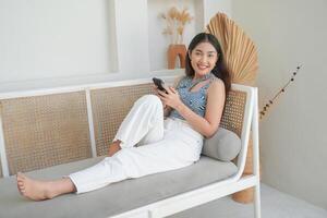 portret van gelukkig Aziatisch vrouw glimlachen en Holding haar smartphone terwijl houdende naar beneden Aan een sofa in villa voor zomer vakantie foto