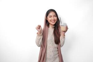 portret van een opgewonden jong Aziatisch vrouw verhogen vuist omhoog gebaar, winnend en vieren zege houding terwijl Holding koffie in een plastic beker. Ramadan en eid mubarak concept foto