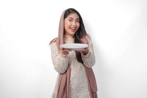 opgewonden Aziatisch moslim vrouw Holding bord en bestek naar eten na vastend terwijl glimlachen vrolijk Aan geïsoleerd wit achtergrond. Ramadan concept foto