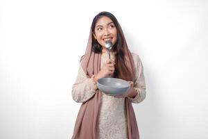 hongerig Aziatisch moslim vrouw Holding kom denken wat naar eten met kopiëren ruimte over- de kom terwijl bijten lepel aan het wachten voor iftar, geïsoleerd wit achtergrond foto