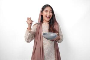 gelukkig Aziatisch moslim vrouw Holding kom en bestek naar eten na vastend terwijl glimlachen vrolijk Aan geïsoleerd wit achtergrond. Ramadan concept foto