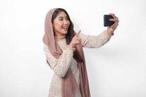 mooi Aziatisch moslim vrouw in hoofddoek hijab sluier lachend en nemen een pret selfie gebruik makend van mobiel telefoon, geïsoleerd door wit achtergrond foto