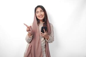 gelukkig Aziatisch moslim vrouw in sluier hijab Holding een slim telefoon terwijl richten naar de kopiëren ruimte Aan de kant over- geïsoleerd wit achtergrond. Ramadan concept foto