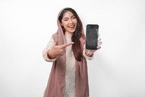 opgewonden jong Aziatisch moslim vrouw in hoofddoek sluier hijab richten naar de kopiëren ruimte Bij smartphone blanco scherm, geïsoleerd Aan wit achtergrond studio. Ramadan en eid mubarak concept. foto