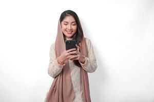 jong Aziatisch moslim vrouw in hoofddoek sluier hijab glimlachen terwijl Holding een smartphone, staand over- geïsoleerd wit achtergrond. Ramadan concept. foto