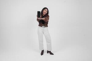 jong Aziatisch vrouw glimlachen breed op zoek gelukkig en Holding smartphone terwijl vervelend gewoontjes kleding staand Aan geïsoleerd wit achtergrond foto
