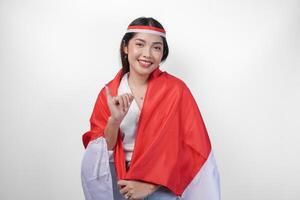 opgewonden Indonesisch vrouw trots tonen weinig vinger gedoopt in Purper inkt na stemmen voor president en parlement verkiezing, vervelend mini vlag hoofdband en wit shirt, aan het bedekken vlag Aan de terug foto