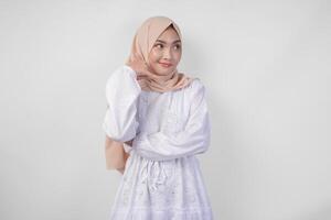 jong Aziatisch moslim vrouw vervelend wit jurk en hijab glimlachen met gelukkig uitdrukking terwijl verhogen hand- naar Doen plukken omhoog telefoon gebaar foto