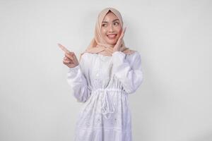 opgewonden Aziatisch vrouw vervelend hijab en wit jurk richten naar de kopiëren ruimte Aan haar Rechtsaf kant, staand over- geïsoleerd wit achtergrond. Ramadan en eid mubarak advertentie concept foto