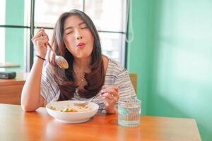 portret van een opgewonden jong Aziatisch vrouw vervelend jurk zittend Bij een restaurant, aan het eten en genieten van ontbijt met een vrolijk glimlach foto
