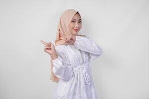 glimlachen Aziatisch vrouw vervelend hijab en wit jurk richten omhoog naar de kopiëren ruimte Aan haar Rechtsaf kant, staand over- geïsoleerd wit achtergrond. Ramadan en eid mubarak advertentie concept foto