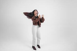 actief Aziatisch meisje is jumping en verhogen handen omhoog slijtage bruin jasje, geïsoleerd wit kleur achtergrond foto