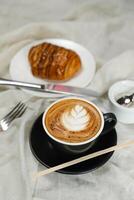 heet macchiato koffie latte kunst geserveerd in kop met croissant, bladerdeeg gebakje, brood en mes geïsoleerd Aan servet kant visie cafe ontbijt foto