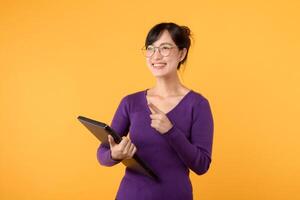 gelukkig Aziatisch vrouw college leerling of leraar gelukkig glimlachen Aan geel achtergrond, Holding laptop. jeugd meisje leerling richten omhoog naar kopiëren ruimte. onderwijs uitwisseling leerling concept foto