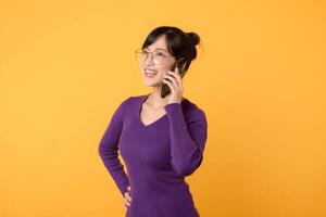 levendig Aziatisch vrouw in haar jaren '30, vervelend Purper overhemd en zonnebril, gebruik makend van smartphone Aan levendig geel achtergrond. nieuw mobiel app concept. foto