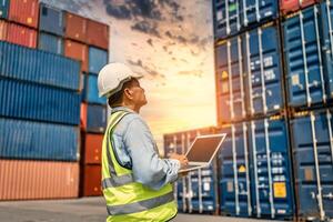 ingenieur of voorman Holding laptop en draagt ppe controle houder opslagruimte met lading houder achtergrond Bij zonsondergang. logistiek globaal importeren of exporteren Verzending industrieel concept. foto