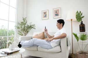 gelukkig Aziatisch Mens zittend Aan bankstel gebruik makend van smartphone in leven kamer Bij huis, kom tot rust tijd en levensstijl concept foto