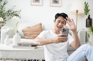 gelukkig Aziatisch Mens zittend Aan bankstel gebruik makend van smartphone in leven kamer Bij huis, kom tot rust tijd en levensstijl concept foto