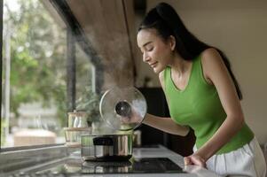 Aziatisch vrouw Koken en ruiken proeverij soep in een pot in de keuken tafel Bij huis. foto