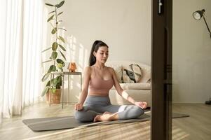 Aziatisch vrouw in sportkleding oefenen en aan het doen yoga in leven kamer Bij huis, gezond levensstijl, mentaal Gezondheid concept. foto