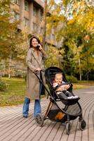 jong vrouw met schattig baby meisje in baby wandelwagen gebruik makend van mobiel telefoon Bij de herfst park foto