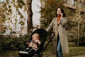 jong vrouw met schattig baby meisje in baby wandelwagen gebruik makend van mobiel telefoon Bij de herfst park foto