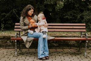 jong vrouw zittend Aan een bank met schattig baby meisje in de herfst park foto